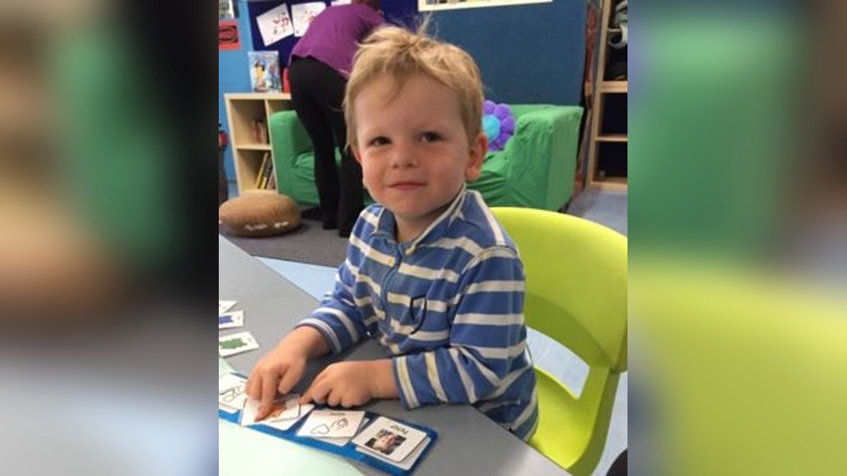 A young boy sitting at a table in an early learning room, smiling, with picture communication cards on a board in front of him