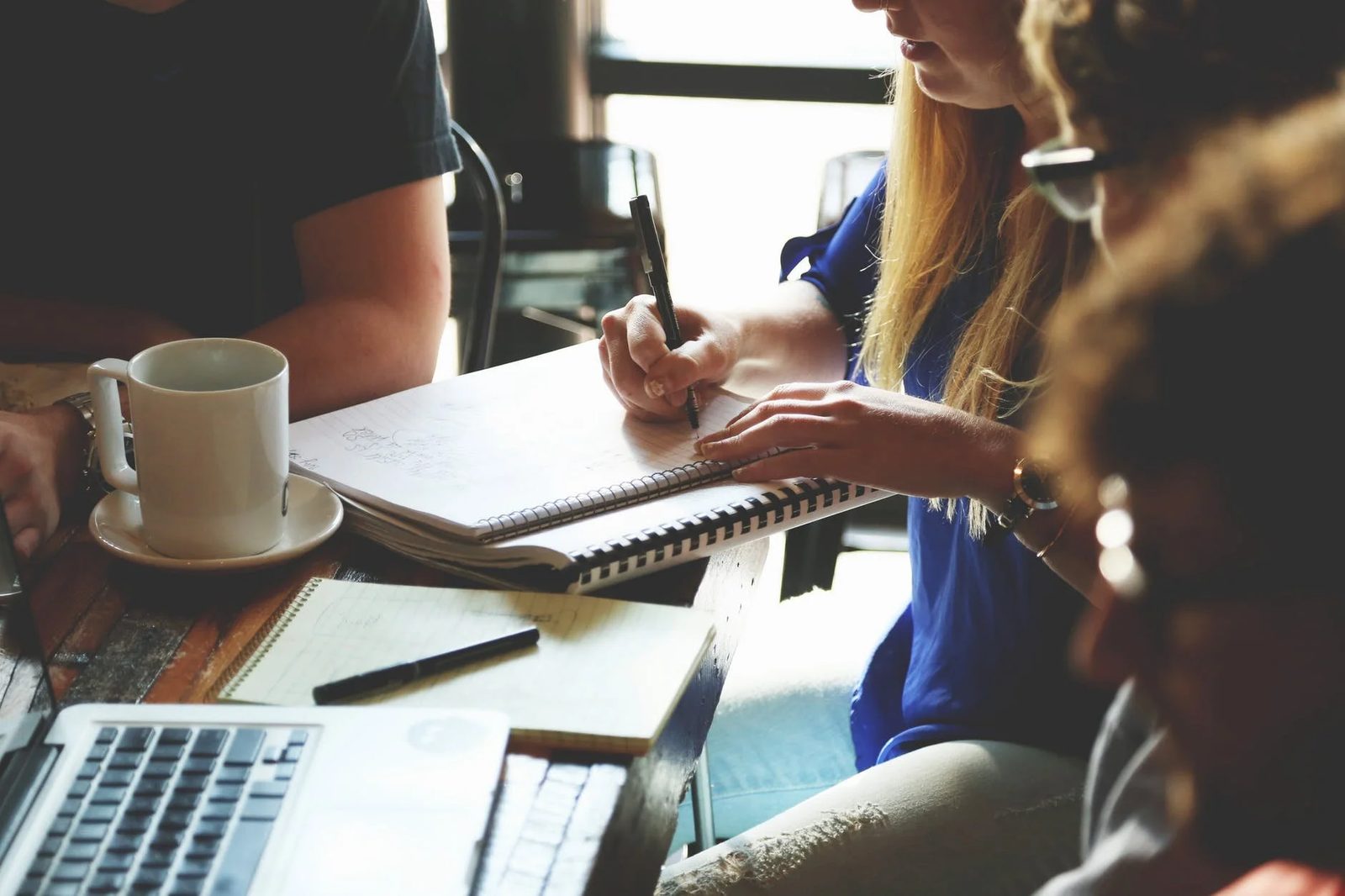 Three people around a café table with notebooks, a laptop and a coffee cup, one of them writing notes.